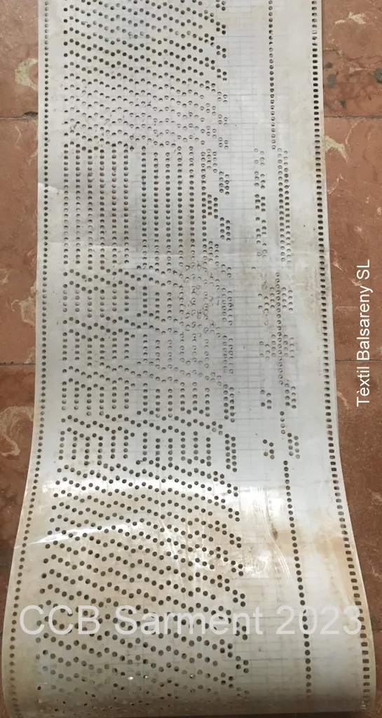 Tira de cartón con filas de perforaciones que codifican el dibujo del tejido